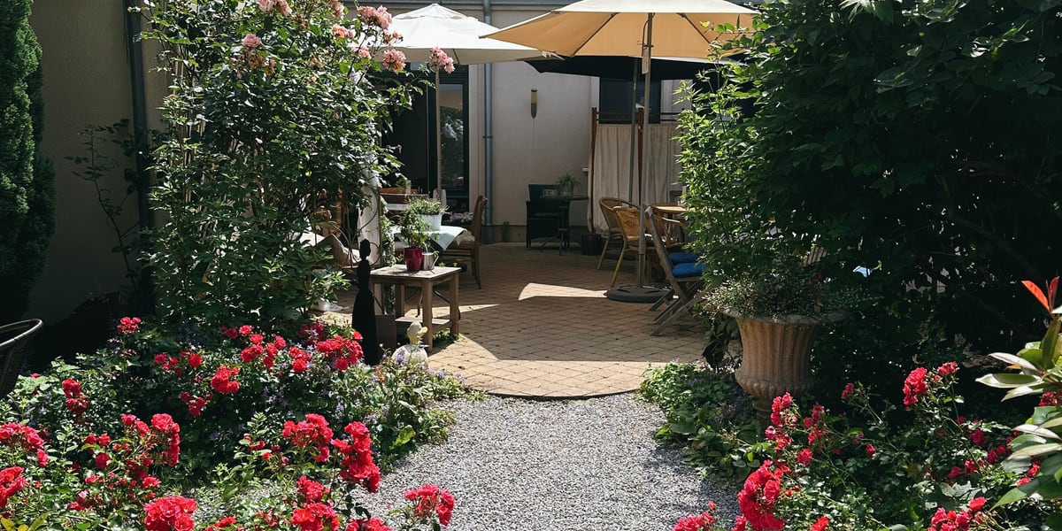 Vom Landschaftsgärtner angelegter Kiesweg zwischen Rosenbeeten führt zu einer überdachten Terrasse mit Sonnenschirmen